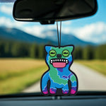 Green-and-blue Fuggler cat-like air freshener with tongue out hanging from a car mirror against a blurred highway and treeline background.
