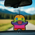 Rainbow-striped Fuggler air freshener with button eyes and open mouth suspended from a rearview mirror with a scenic blurred road behind.
