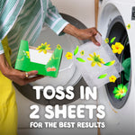 Hands holding a Gain box and placing a dryer sheet into a front-loading washer with text ‘TOSS IN 2 SHEETS’