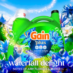 Close-up of a green Gain Plus detergent jug with blue floral label surrounded by bubbles and blue flowers, 'waterfall delight' text