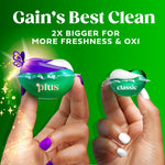 Two Gain detergent pacs held between fingers comparing larger 'plus' pac to smaller 'classic' on green background