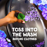 Hand pouring purple scent beads from a green measuring cup into a washing machine drum with text instructing to toss into the wash