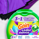 Partial top view of a Gain green tub next to folded athletic clothing, showing '3 in 1 + flings! Odor Eliminators' label