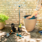 Outdoor scene of a coiled garden hose being placed into a tall planter beside gardening tools and a brick wall