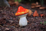Small orange and yellow-tinted spotted mushroom figurine set in garden mulch and dried leaves.
