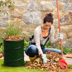 Person raking leaves next to a green collapsible pop-up trash can filled with yard debris in a garden setting