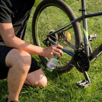 Lifestyle photo of a person kneeling on grass spraying Gear Hugger degreaser at a bicycle drivetrain near the rear wheel.