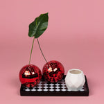 Small white faceted ceramic planter styled on a black-and-white tray with red disco-ball props on pink background