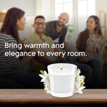 Lifestyle image with a lit Glade clear-jar candle and white flowers on a table in front of a blurred group of people and text about warmth and elegance