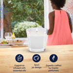 Lifestyle image showing a lit Glade jar candle on a wooden surface with blurred window view and a person in coral dress in background