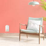 Minimal styled room with a Glade PlugIns warmer mounted low on a coral-colored wall beside a mid-century chair and hanging lamp