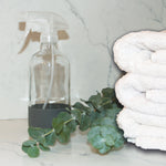 Clear glass spray bottle with gray rubber base styled on a marble surface beside eucalyptus and towels