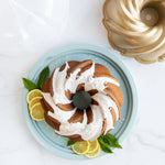 Top-down styled shot of a glazed bundt cake on a teal base with gold bundt pan and clear lid parts nearby