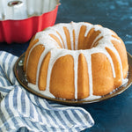 Glazed bundt cake on a plate with icing drips, a striped towel, and a red bundt pan visible in the background.