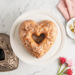 Glazed floral heart bundt cake on a white platter with part of the metallic floral heart pan visible at the edge and table styling.