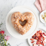Glazed heart-shaped bundt cake on a white plate, styled with strawberries and flowers, made from a floral heart mold.