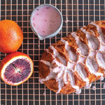Iced loaf cake with geometric pattern on a cooling rack, accompanied by whole and halved blood oranges and a small pitcher of glaze.