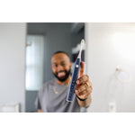 Person holding a navy Gleem electric toothbrush toward the camera with bathroom background blurred