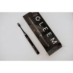 Black Gleem toothbrush laid next to its black retail box on a light speckled surface in a flat lay composition.