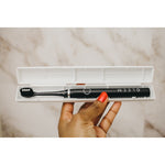 Hand holding an open white travel case containing a black Gleem electric toothbrush, shown over a neutral background.