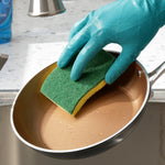 Hand wearing a teal glove using a green-and-yellow scrub sponge to clean a nonstick frying pan under a faucet