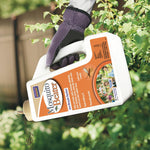 Gloved hand holding a tilted Bonide Mosquito Beater jug over garden foliage, label visible