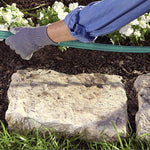 Gloved hand holding a green soaker hose laid along garden soil and next to flowering plants and stepping stones