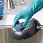Gloved hand holding a blue-gray steel wool soap pad while scrubbing the exterior of a frying pan over a sink