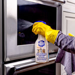 Person wearing yellow gloves spraying Bar Keepers Friend stainless steel cleaner at a stainless oven door in a kitchen scene.