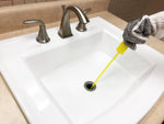 Yellow barbed drain-cleaning stick inserted into a bathroom sink drain held by a gloved hand