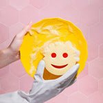 Hands wearing white gloves holding a yellow plate being cleaned by a pale yellow smile-face Scrub Daddy sponge