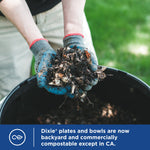 Close-up of gloved hands holding a handful of compostable organic material over a compost bin with a denim-clad person in background.