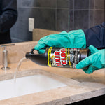 Gloved hands pouring Instant Power Hair Clog Remover from a tilted bottle into a bathroom sink drain