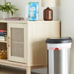 Lifestyle scene showing a Great Value box of medium drawstring trash bags on a wooden cabinet next to a stainless step trash can and lamp.