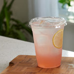 Clear plastic cup with sip lid filled with iced pink lemonade and a lemon slice on a wooden board in a kitchen setting