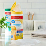 Open yellow canister of Great Value disinfecting wipes on a bathroom counter with a pulled wipe and another canister blurred behind
