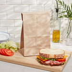 Self-standing brown paper lunch bag staged on a cutting board next to a sandwich and tomatoes in a kitchen setting.