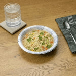 Disposable patterned paper bowl on a wood table filled with noodle soup, shown beside a glass of water and utensils.