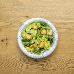 Top-down view of a decorative disposable paper bowl on a wood table filled with green salad and croutons.