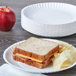Styled scene with stacked white paper plates and a sandwich with chips on a plate, apple to the side on a wooden surface