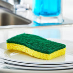 Close-up lifestyle shot of a green scouring pad on yellow sponge resting on stacked white plates near a sink