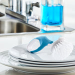 Blue-handled round-bristle scrubber resting on stacked white plates with kitchen sink and soap dispenser blurred in background