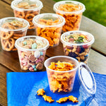 Several clear snack cups with snap-on lids filled with various snacks arranged on a wooden table, one lid removed.