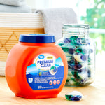 Lifestyle shot of the orange Great Value Premium Clean tub beside a glass jar filled with blue-green laundry pacs on a wooden surface