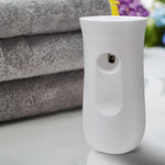 White automatic air freshener dispenser photographed on a surface with folded towels and blurred flowers in the background.