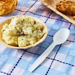 White plastic disposable spoon placed beside a bowl of mashed potatoes on a blue checkered tablecloth