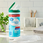 Great Value disinfecting wipes container on a bathroom countertop with lid open and a wipe pulled out, toothbrushes and plants in background