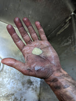Grimy work hand holding a small dollop of light-colored hand cleaner in the palm over a metal sink