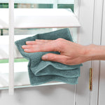 Hand cleaning white window blinds with a teal microfiber cloth, showing cloth texture and cleaning action