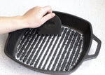 Hand using a black Lodge multi-tooth scraper inside a ridged cast-iron grill pan to clear residue, logo visible.